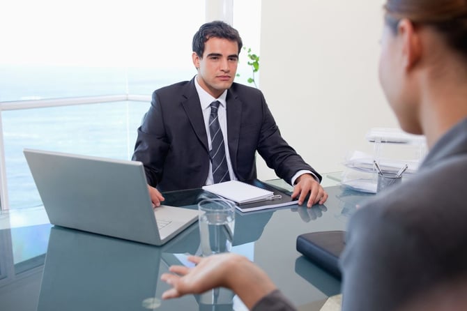 A manager listens attentively during a one-on-one meeting with an employee across a glass conference table, with a laptop and notebook nearby.