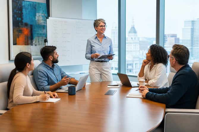 A woman stands and presents to five business professionals seated around a wood conference table, a whiteboard of notes behind her and a city skyline through the windows, during a facilitated peer group discussion.