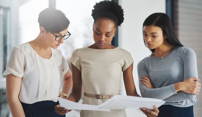 Three business owners stand in a bright office and review a one-page decision summary together, pressure-testing a high-stakes call with outside input.