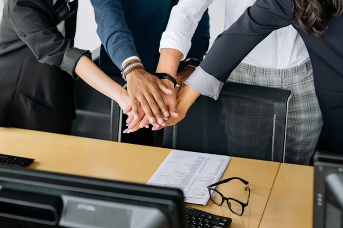 Business team stacking hands over a conference table in a show of alignment, with a printed plan and glasses on the desk