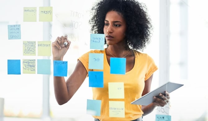 A woman holding a tablet writes on a glass wall covered in blue and yellow sticky notes while mapping out a strategy