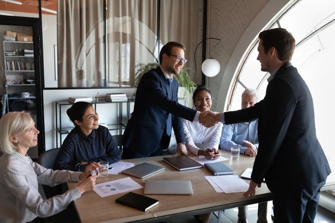 A business leader shakes hands with a newly promoted manager across a conference table while smiling colleagues look on, representing a formal internal promotion moment.