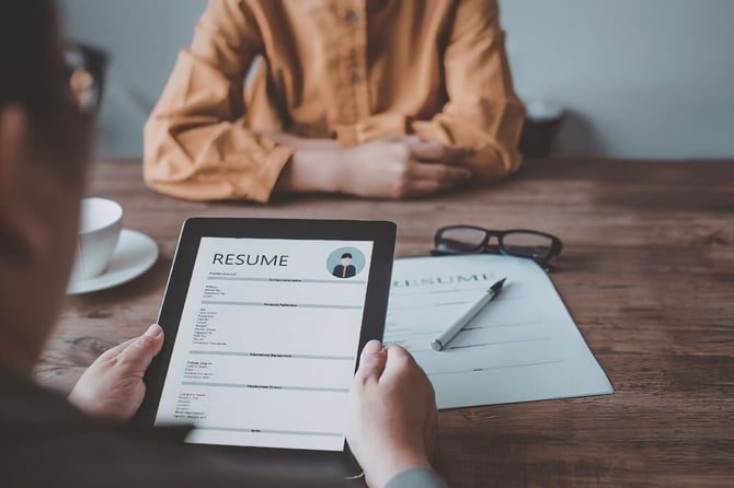 A hiring manager holds a tablet displaying a candidate's resume during a face-to-face job interview, with a printed resume and pen on the table nearby