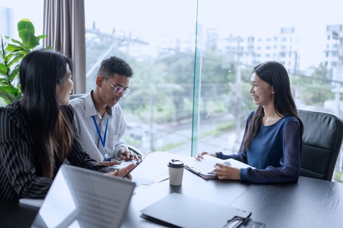Two hiring managers review notes and interview a smiling job candidate across a conference table in a bright, modern office
