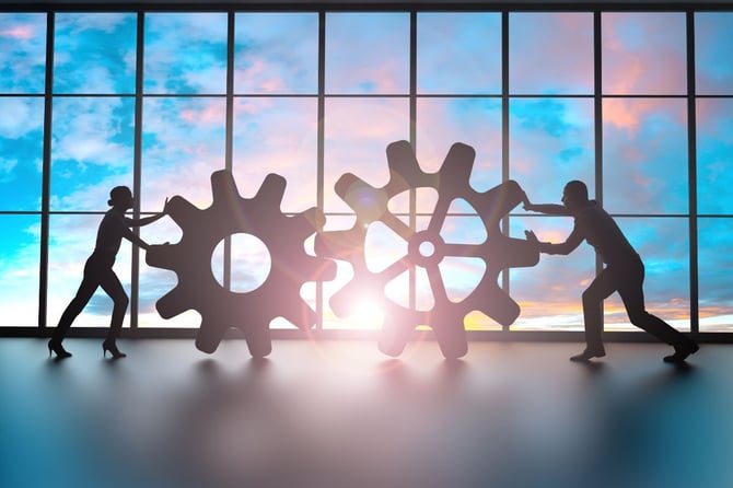 Two business professionals push large interlocking gears together in a sunlit office, representing teamwork and the mechanics of building a strong company culture in a small business.