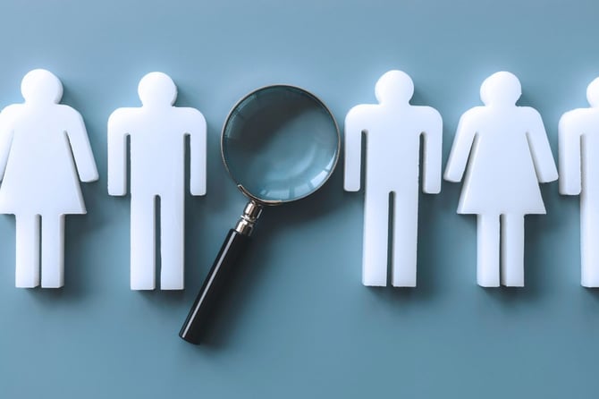 White paper cutout figures standing in a row with a magnifying glass highlighting one, representing the employee screening and selection process