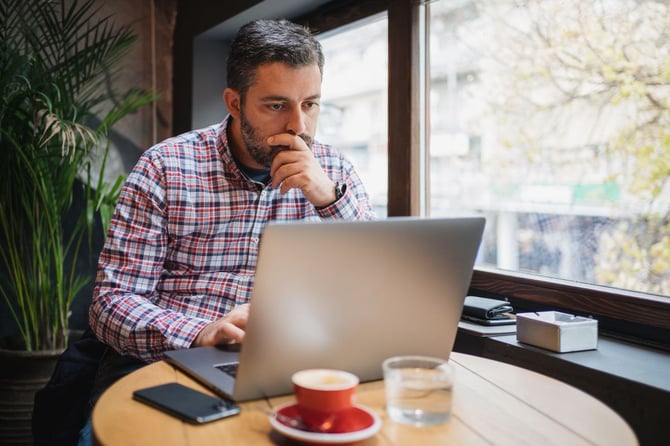 A business owner sits alone at a café table, looking pensively out the window while working on a laptop.