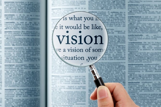 A magnifying glass held over an open dictionary page enlarges the word "vision" and part of its definition, with the surrounding dictionary text blurred in the background.