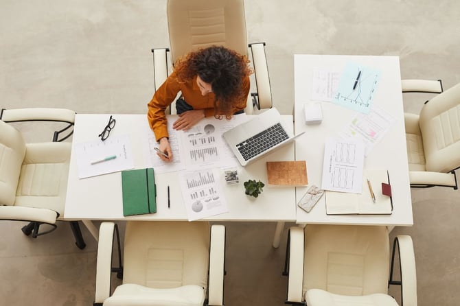 A manager reviews performance reports and charts spread across a conference table, with a laptop open and notebooks nearby, seen from a bird's-eye view.
