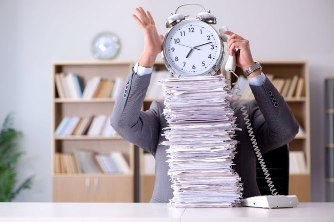 A businessperson sitting at a desk behind an overwhelming stack of papers, with a large alarm clock obscuring their face and one hand raised in frustration, illustrating the busyness and time pressure that prevents manager development.