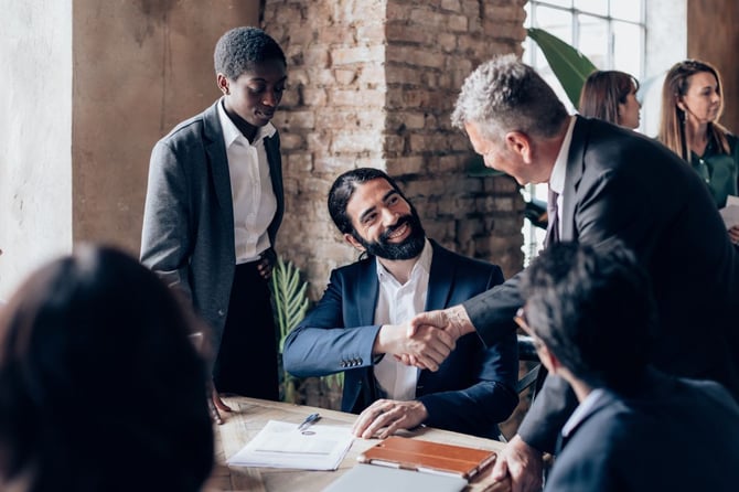 A smiling manager is welcomed by the team during his first day, exchanging a handshake in a collaborative office setting.